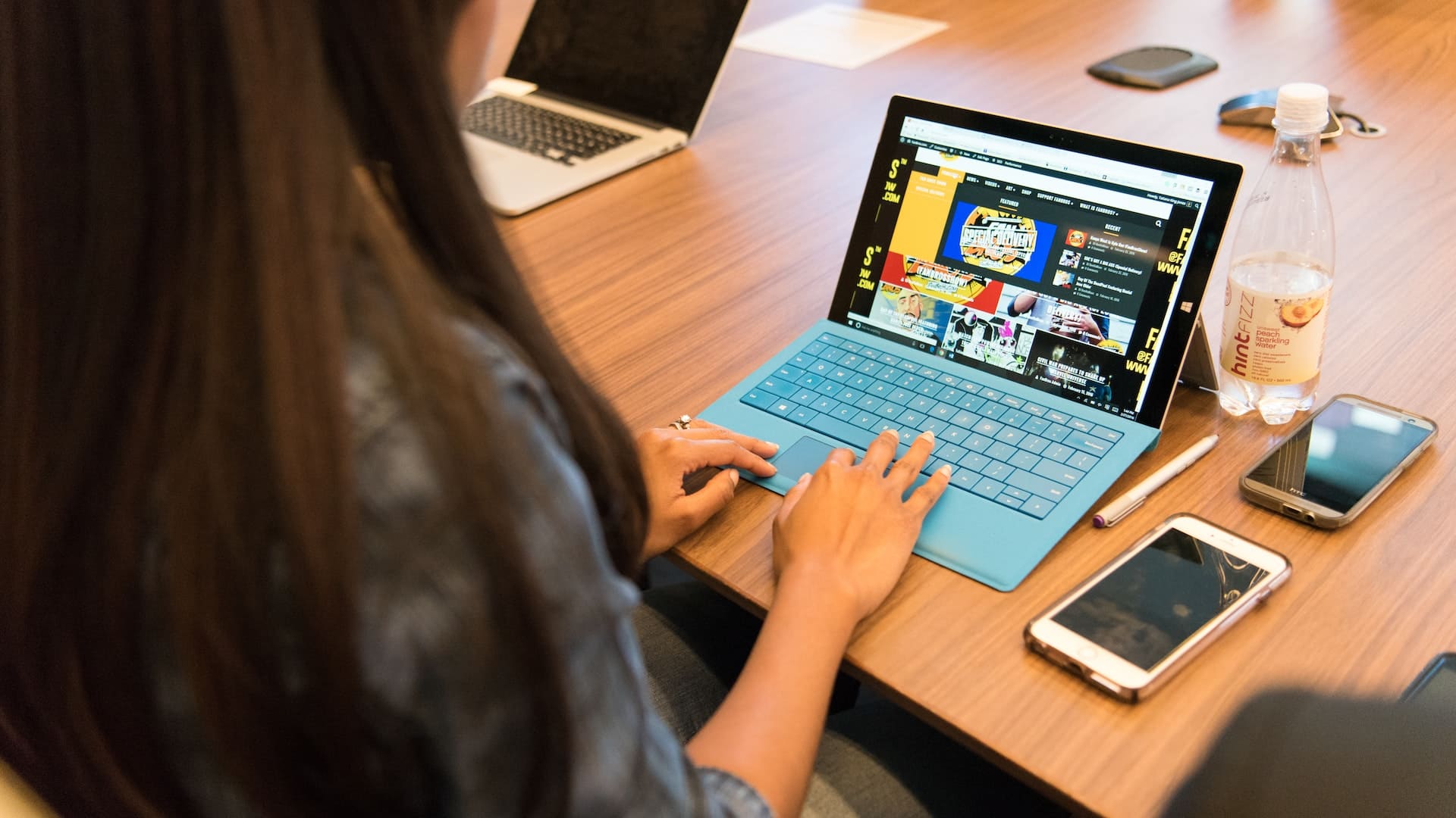 Mulher web designer sentada usa computador em mesa de madeira. Há também um celular e outros objetos de escritório sobre a mesa.