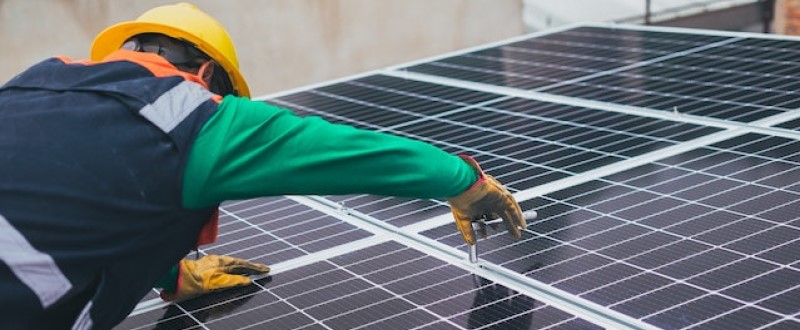 Técnico em energia solar, equipado com capacete e luvas amarelas, instala painéis fotovoltáicos em uma superfície inclinada.