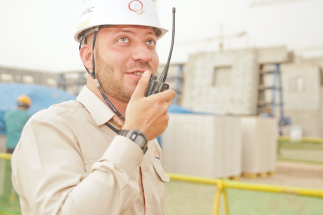 Homem, engenheiro civil, com capacete e rádiotransmissor na mão. Peças de construção pré-moldadas ao fundo.