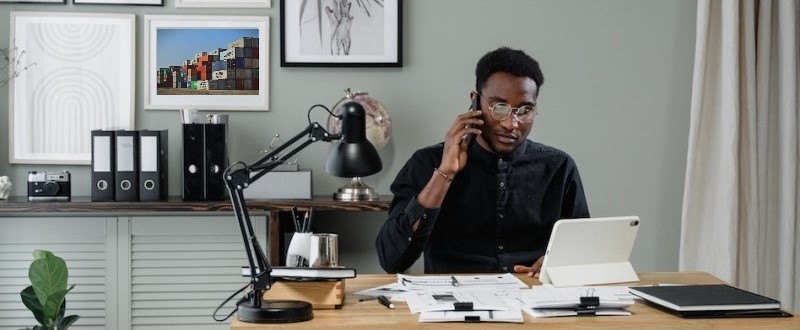 Homem negro, em um escritório, fala ao telefone olhando para a tela de um computador. Na parede há um quadro com uma foto de contêineres em um porto.