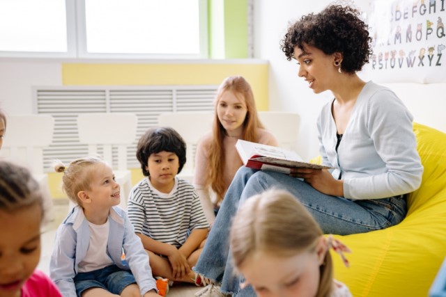 Professora de ensino fundamental, sentada, lendo livro em sala de aula para um grupo de cinco crianças de diferentes etnias.