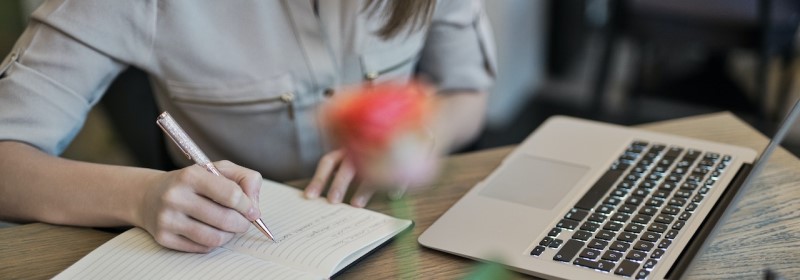 Mulher com caneta na mão faz anotação em caderno sobre a mesa, em frente a um computador.