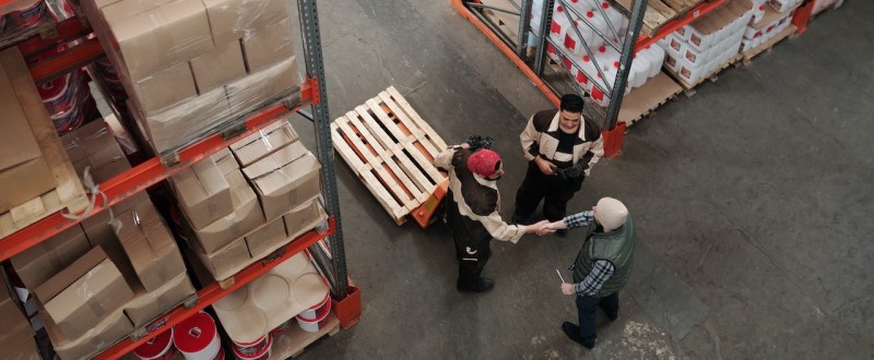 Vista de cima de um supervisor de distribuição e dois outros homens conversando entre prateleiras em um depósito com estantes cheias de caixas de produtos.