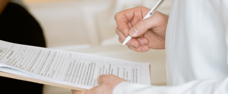 Foto de mãos de um médico segurando uma prancheta com questionário e fazendo anotações.