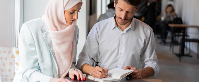 Homem e branco escrevendo em um caderno ao lado de uma mulher com lenço na cabeça.